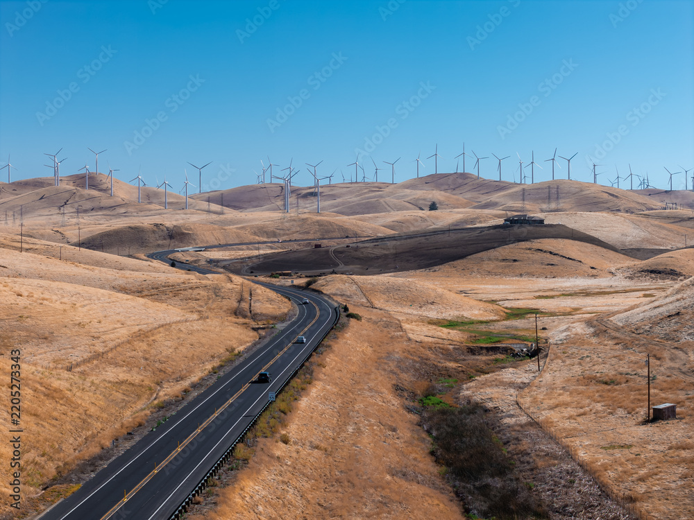 Fototapeta premium Aerial view shows vehicles driving along a winding asphalt highway through arid rolling hills, leading toward a large wind farm with spinning turbines in California.