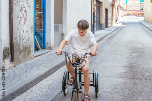 Cuadro en lienzo Young man with Down syndrome riding adult tricycle alone through street in Spani