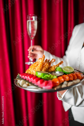 A server holding a platter of fried shrimp and sliced sashimi alongside a glass of champagne against a red curtain.