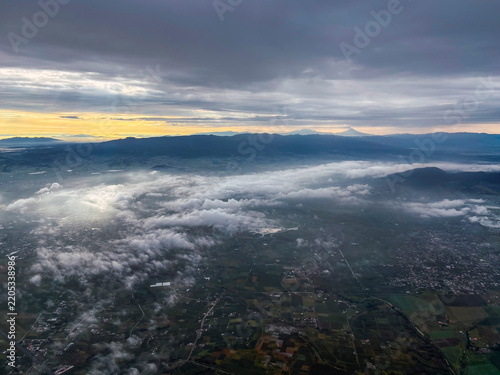 Aerial photograph of a landscape in Ixtlahuaca, Mexico, at sunrise, featuring clouds and a sky with very warm tones; Santa Cruz Hill is visible, with Popo and Izta in the background.
