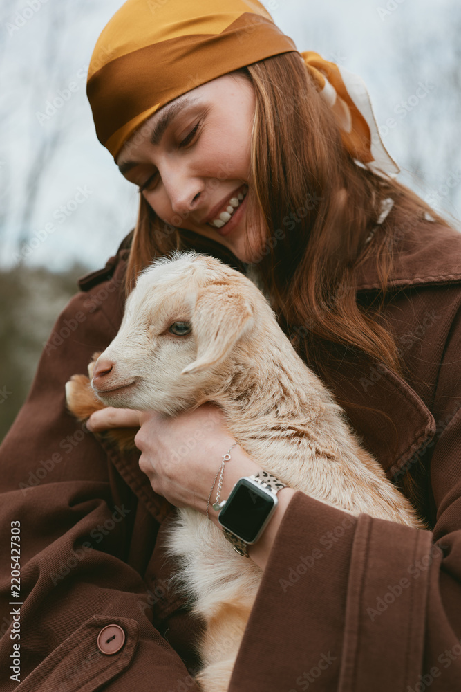 Obraz premium Joyful woman in autumn coat holds a young goat kid close, sharing affectionate moment outdoors, warm scarf, smile, gentle interaction, rustic mood, lifestyle scene, candid portrait, natural light,