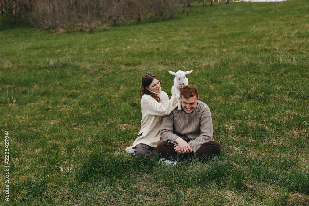 Obraz premium Happy couple sits on a grassy field with a playful goat kid between them, sharing a gentle moment in cozy sweaters, natural light, candid lifestyle portrait outdoors, loving interaction and rural