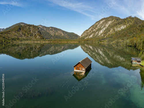 Aerial view of a wooden boathouse floating on the calm, clear waters of Attensee, surrounded by forested mountains under a bright blue sky in Rottenbach, Bayern, Germany.