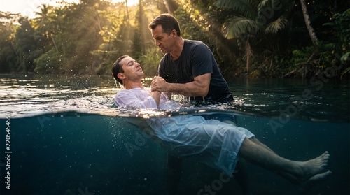Man receives baptism in a tropical river surrounded by lush green jungle foliage at sunset