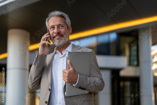 Confident mature businessman talking on phone holding laptop
