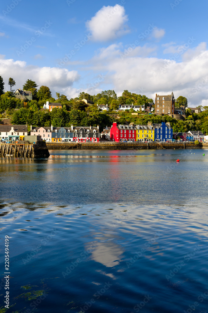 Fototapeta premium Colorful painted houses along Tobermory harbor waterfront, Scotland, calm blue sea water reflecting bright facades under wooded hillside