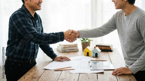 Two men shaking hands over a wooden table with architectural blueprints and small house models to finalize a successful real estate property deal