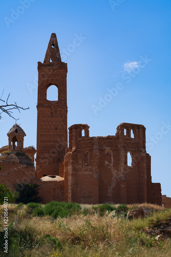 An ancient ruined bell tower rises above collapsed walls of a deserted settlement in dry grassland. Ornate brickwork and pointed arches showcase the faded grandeur of this crumbling abandoned landmark