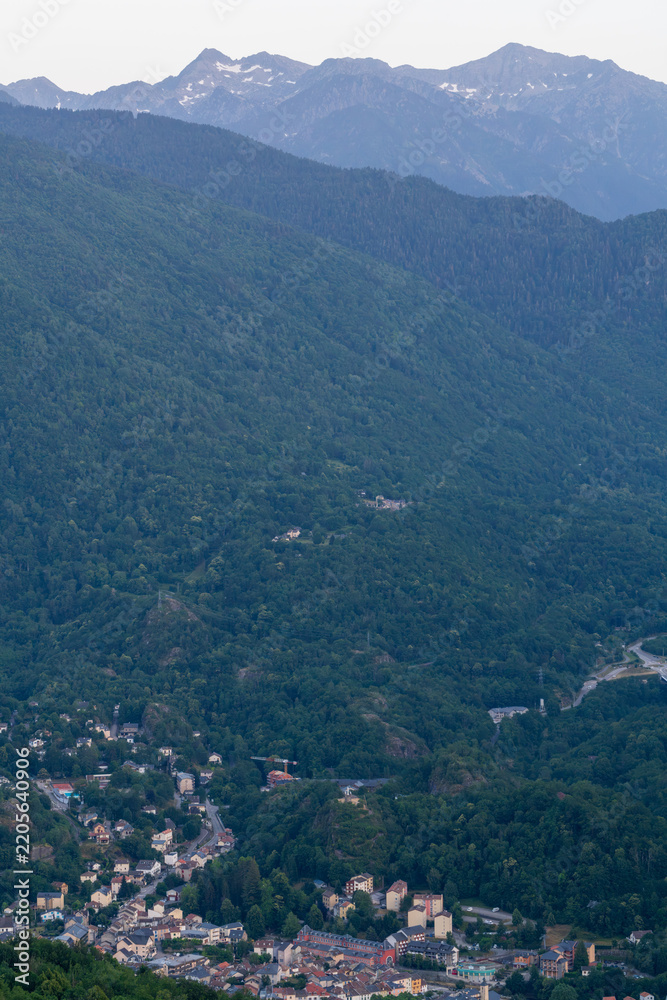Naklejka premium Houses cluster together at the foot of a lush, wooded mountainside in a deep alpine valley, below rugged terrain. Snowy distant peaks loom above the dense forest canopy beneath a soft hazy evening sky