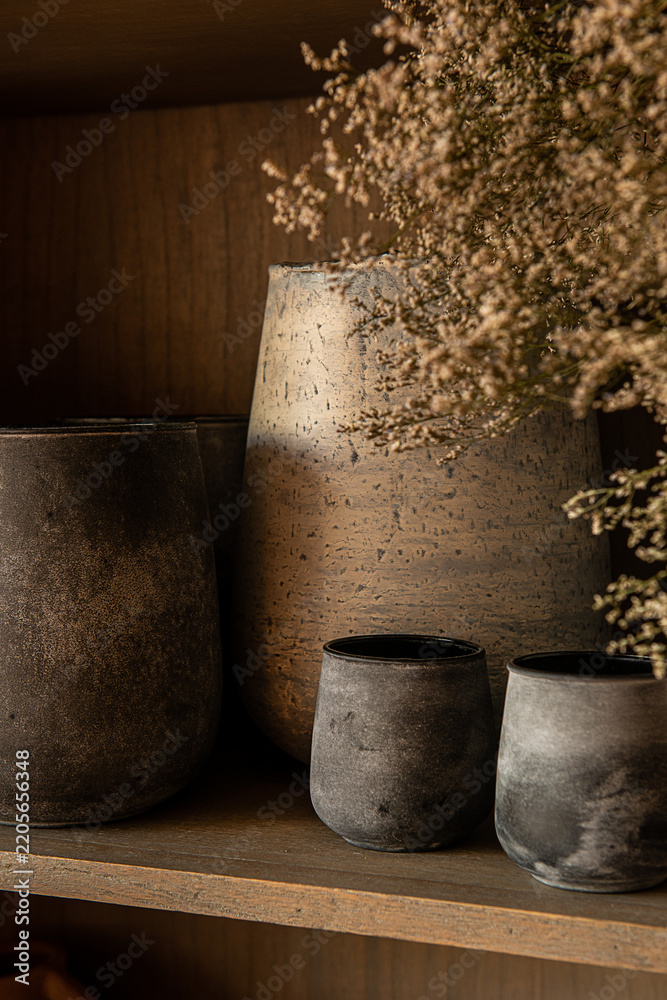 Fototapeta premium Close up of rustic ceramic cups and textured earthenware vase filled with delicate dried flowers on a wooden shelf.