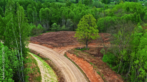Aerial view of a winding dirt road cutting through a lush green forest with a recently cleared patch of land in Latvija.
