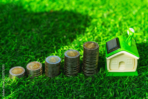 Green energy home and eco housing investment concept, miniature house with solar panel and wind turbine beside rising coin stacks on lawn, symbolizing renewable savings and sustainable value.