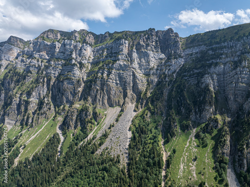 Aerial view of the dramatic limestone cliffs and steep scree slopes of the Justistal valley near the Niederhorn, with lush alpine forests and meadows in Sigriswil, Bern, Switzerland.