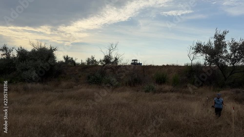 Deminer in protective vest and visor helmet walking through a dry grass field marked with stakes during humanitarian mine clearance