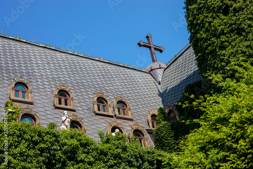 Historic church roof with dark slate tiles and tall stone cross standing above covered in lush green ivy vines, gothic architecture set against clear blue sky backdrop, high quality photo