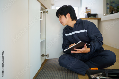 A focused young Asian repairman crouches on the floor examining near open kitchen cabinet with clipboard in hand, ideal for home maintenance and service concepts