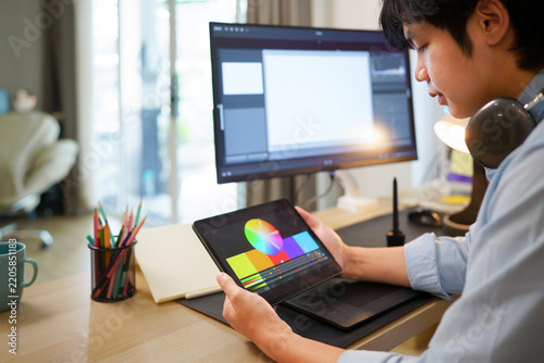 Young Asian graphic designer reviewing color wheel palette on tablet at creative workstation with monitor and design tools, ideal for creative and tech concepts