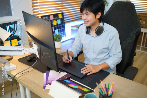 Young Asian male graphic designer working on drawing tablet with stylus pen and monitor in a modern home studio with color swatches