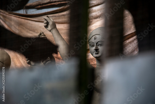 A detailed close-up showing the raw, unpainted clay surface and fine features of an unfinished Durga idol face, captured through the iron security grill of an artisan workshop in Kumartuli, Kolkata. 