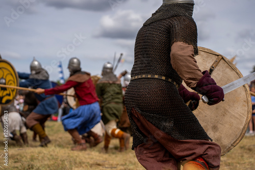 Fotografie Historical reenactment of medieval battle with warriors wearing chainmail armor and helmets while fighting with swords and round wooden shields outdoors