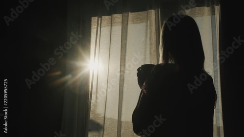 Silhouette of woman drinking coffee by window during sunrise with lens flare effect.