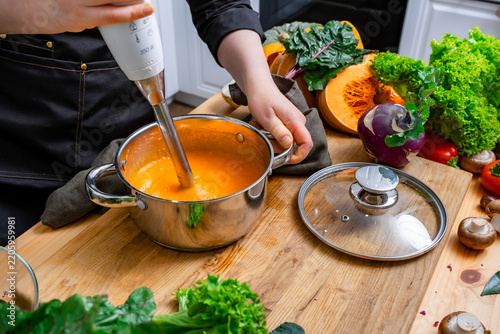 Comfort autumn food — chef hands blending hot pumpkin soup in metal pot. High haute cuisine meal preparation using modern kitchen appliance. Healthy eating lifestyle with seasonal organic dish.
