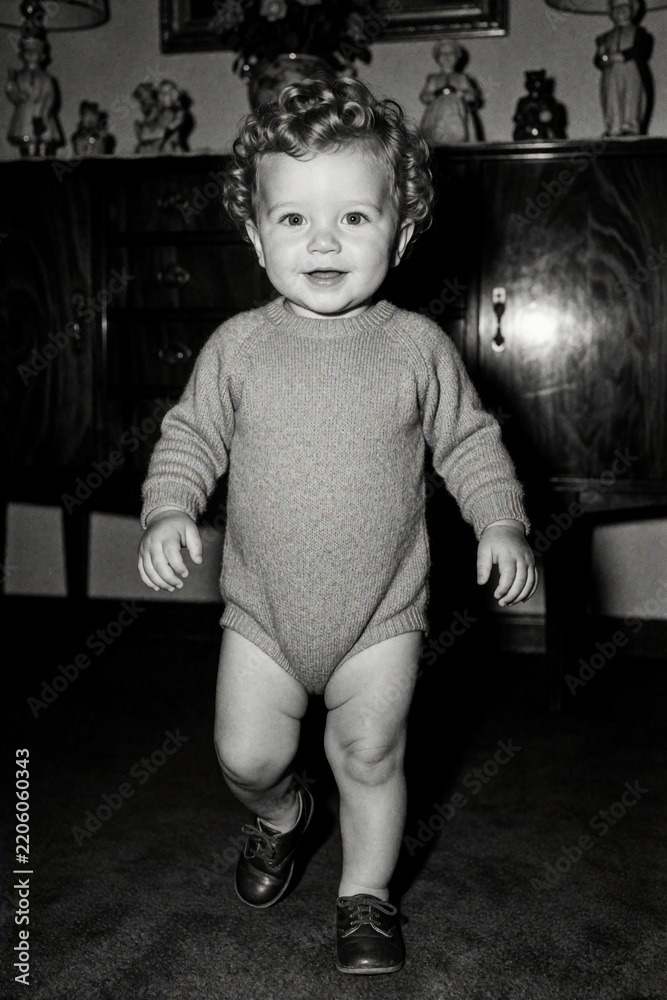 Fototapeta premium Vintage black, white portrait of cute curly haired toddler boy. Small child learns to walk alone in retro home living room. Happy little boy wears knit romper, small shoes during childhood milestone.