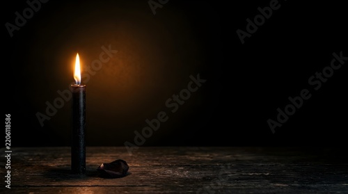 Single black taper candle burning with a bright warm flame on a rustic wooden table beside a dried rose petal on dark background.