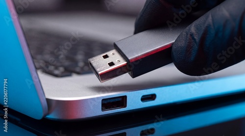 USB malware hacker flash drive laptop cybersecurity data breach attack Gloved hand holds a portable storage device beside a computer port suggesting infection theft or unauthorized file transfer