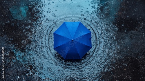 A bright blue umbrella creates ripples in a puddle of rainwater on a reflective surface during a rainy day