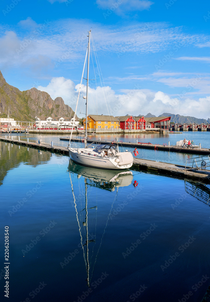 Fototapeta premium Sailboat at a pier in Svolvaer harbor in the Lofoten Islands with calm water, colorful houses and distinctive mountains beneath a clear blue sky.