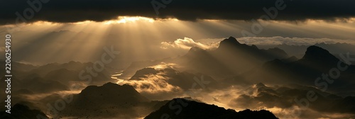 a breathtaking view of a mountainous range bathed in golden light. The suns rays pass through the clouds, creating a radiant glow that contrasts with the dark, stormy sky.