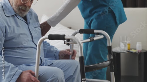Nursing home female worker helping elderly man move with walking frame, rehab