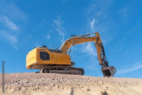 Powerful, yellow excavator on a crest of loose, sandy-colored earth, its tracks firmly embedded in uneven terrain. Boom arm extended upwards. Large metal poised bucket. 