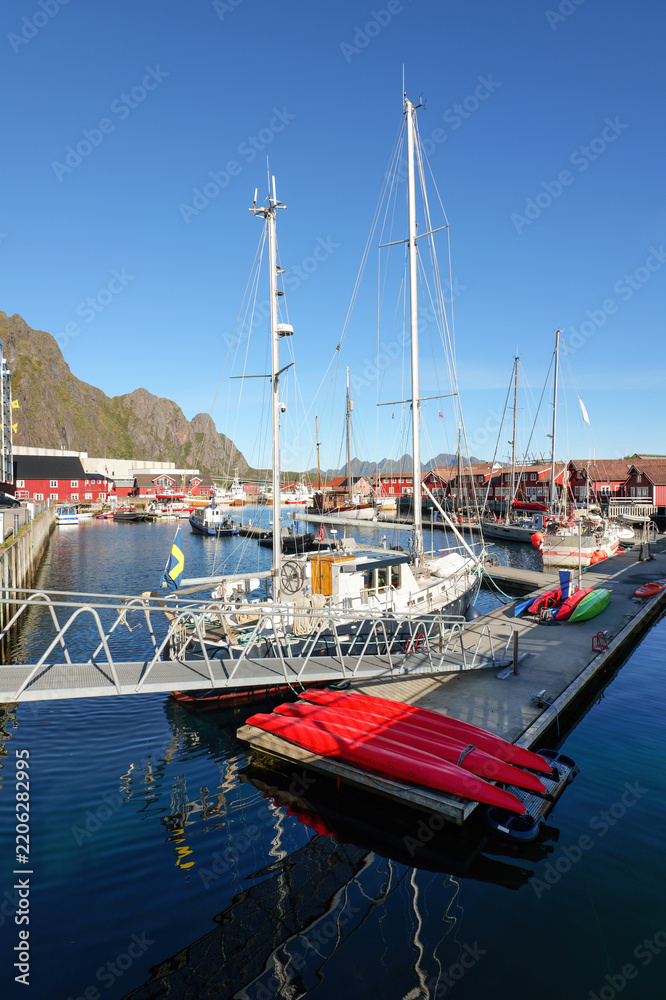 Fototapeta premium Sailboats and colorful kayaks lie in the sunny harbor of Svolvaer in the Lofoten Islands, surrounded by red wooden houses, calm water and rugged mountains.