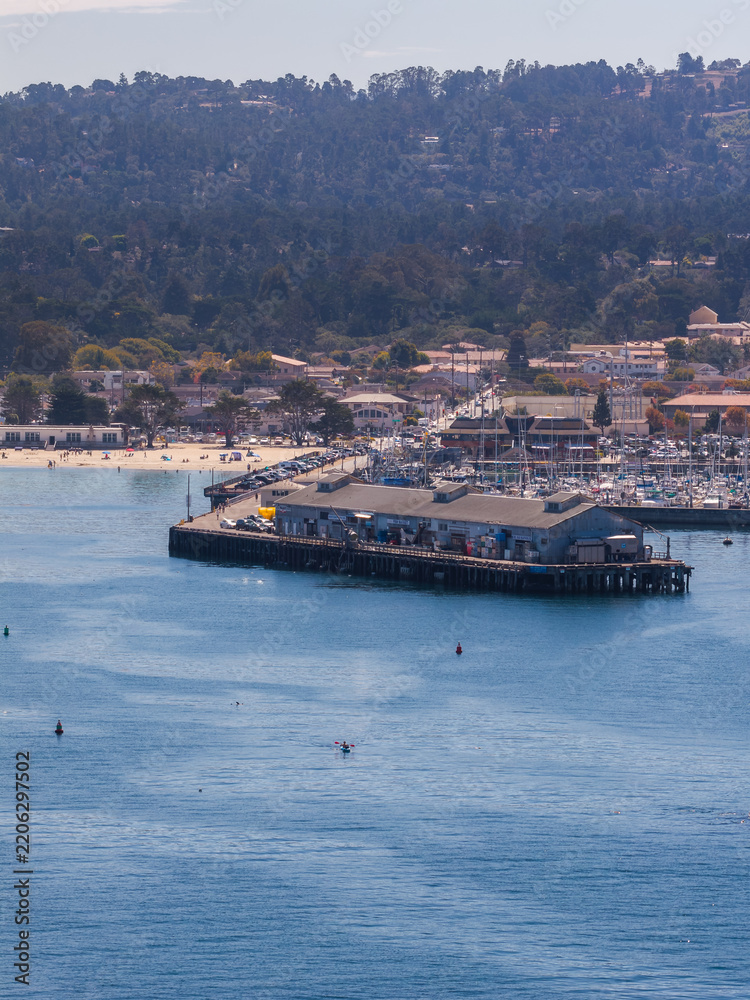 Fototapeta premium Aerial shot reveals the wooden wharf pier in Monterey, California, with kayakers paddling in the bay water, boats moored in the marina, and coastal hills visible.
