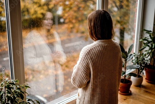 A person stands by a window, looking out at the autumn scenery with fallen leaves. Their reflection is visible in the glass, creating a sense of introspection and quiet contemplation.