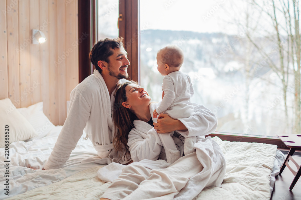Fototapeta premium Happy family playing on bed enjoying snowy mountains landscape from hotel window. Mom and dad having fun with toddler baby girl