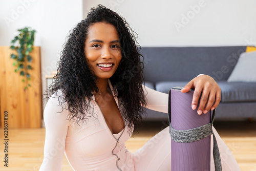 Portrait of a young Latin American woman smiling while holding a rolled yoga mat at home. Happy Colombian millennial female relaxing before fitness training. Healthy lifestyle and fitness concept.
