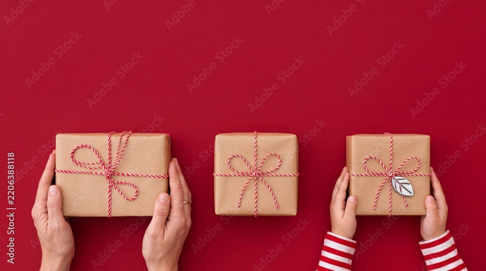 Obraz premium Top view of hands of an adult and child holding Christmas gift boxes wrapped in craft paper with red twine on a vibrant red background. Minimalist holiday preparation and giving presents concept.