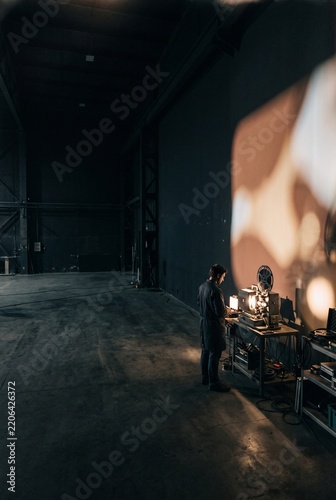 Projectionist operating a vintage 35mm film reel projector in a spacious dark industrial warehouse studio