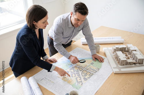 Two professional architects review a city development plan and architectural model on a wooden table in a bright office setting while using a magnifying glass for detail