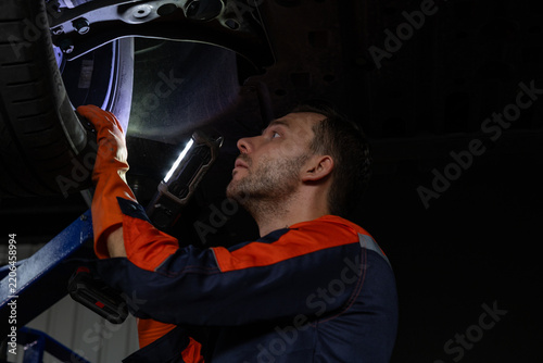 Mechanic in orange protective clothing inspects vehicle suspension inside dark garage using handheld work light with focused attention and safety care