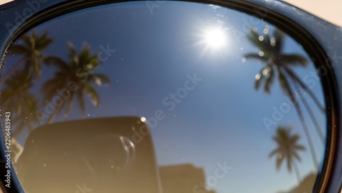 Reflection of palm trees and bright sun in a car rearview mirror under a clear blue tropical sky