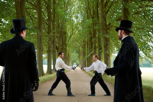 Two Victorian gentlemen engage in a formal sword duel on a gravel path lined with tall trees, while two witnesses in top hats and long coats observe the honorable combat at dawn.