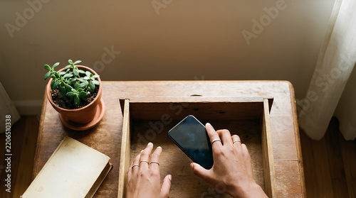 Hands carefully set a smartphone face-down into a wooden drawer beside a book and plant in a calm minimal scene