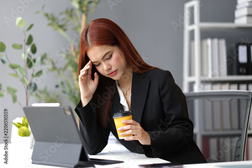 Young Asian businesswoman suffering from headache, holding coffee cup and touching her forehead while sitting at office desk and working with digital tablet.