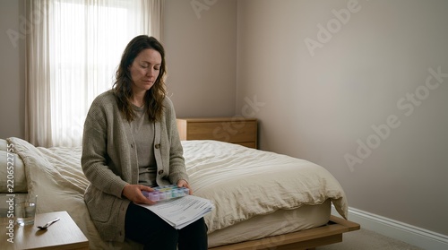 Woman in her thirties with a weary expression organizes medication and paperwork on the edge of a bed in documentary style