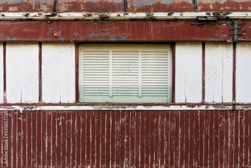 Weathered Wooden Facade With Closed Louvered Window. Aged wooden building exterior with closed shutters, faded red and white paint, peeling surfaces, and rustic architectural texture.
