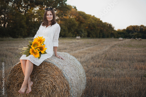 woman in a white dress holding a bouquet of wild flowers with sunflowers in a mowed field with straw bales